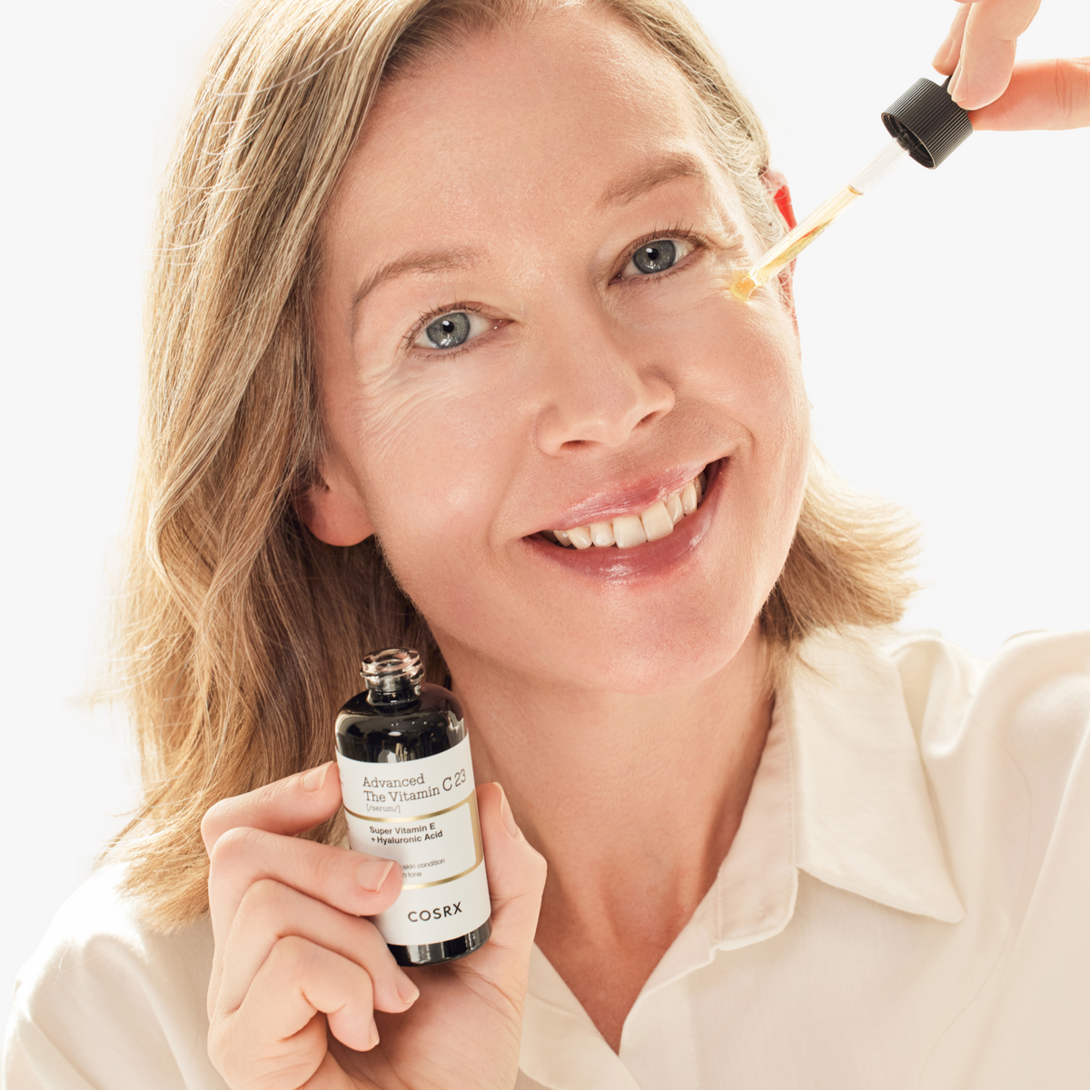 Smiling mature model applying COSRX Advanced The Vitamin C 23 Serum to the under-eye area; clinically proven antioxidant-rich formula enhances elasticity, smooths wrinkles, and brightens uneven tone for visibly youthful skin. Triple-complexion redness improved by 30.14% after 4 weeks of consistent use.