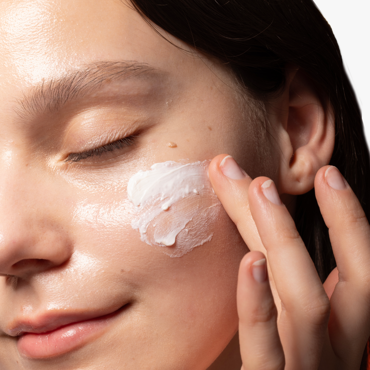 Close-up of model gently applying COSRX Vitamin E Vitalizing Sunscreen SPF 50+ on cheek; creamy vitamin E formula delivering antioxidant-rich moisture and SPF 50+ protection while maintaining a smooth, white-cast-free complexion under makeup.